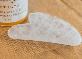 White quartz gua sha stone on a wooden table in front of a bottle of Face Food face oil